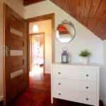 Przytulny korytarz w domku na Kaszubach z drewnianą podłogą i sufitem, białą komodą, lustrem i otwartymi drzwiami