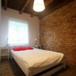 Przytulna sypialnia w domku na Kaszubach z białym łóżkiem, ścianą z czerwonej cegły i sufitem z drewnianymi belkami