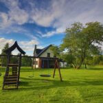 Zielony ogród z drewnianym placem zabaw i dużym drzewem, obok domku na Kaszubach w słoneczny dzień